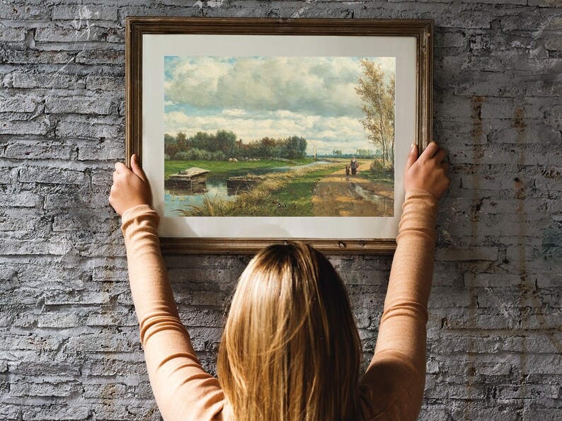 A person hangs a framed landscape painting on a brick wall. The painting depicts a rural scene with a river, trees, and a cloudy sky.