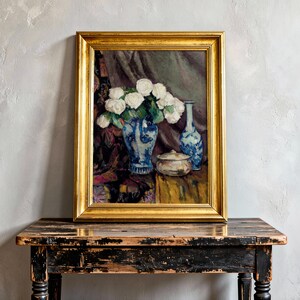 A framed painting of white roses in a blue vase sits on a rustic wooden table against a textured light gray wall, conveying elegance and serenity.