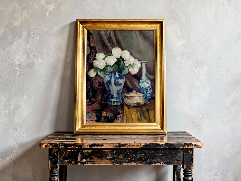 A framed painting of white roses in a blue vase sits on a rustic wooden table against a textured light gray wall, conveying elegance and serenity.