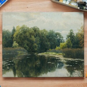 An oil painting depicting a tranquil landscape scene. The artwork showcases a serene lake reflecting lush green trees and foliage under a cloudy sky. Paintbrushes and art supplies surround the painting, suggesting an artist's workspace.
