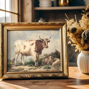 Ornate framed painting of a cow and sheep in a rustic scene, sits on a wooden table near a window. A vase with dried flowers adds warmth and charm.