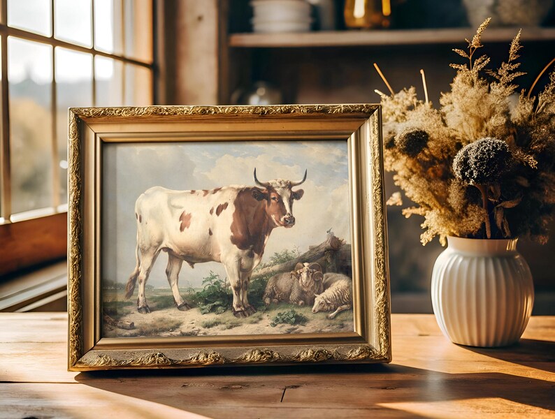 Ornate framed painting of a cow and sheep in a rustic scene, sits on a wooden table near a window. A vase with dried flowers adds warmth and charm.