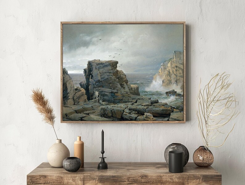 Framed seascape painting on a wall above a wooden shelf. The scene shows rugged cliffs and waves, with decor items like vases and candles below.