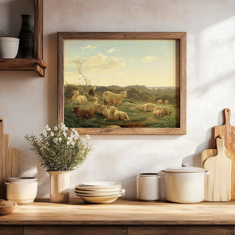 Framed painting of sheep in a serene landscape hangs on a kitchen wall. Below, a wooden counter holds white dishes, pots, and a vase with flowers.