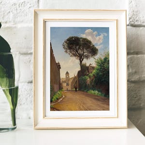 Framed painting on a white shelf shows a sunlit, tree-lined street leading to a tower, with a person walking. A leafy plant in a glass sits nearby, set against a brick wall.