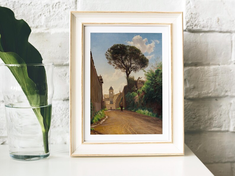 Framed painting on a white shelf shows a sunlit, tree-lined street leading to a tower, with a person walking. A leafy plant in a glass sits nearby, set against a brick wall.