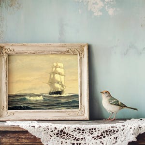 A framed painting of a tall sailing ship on the ocean, with billowing sails and waves, placed on a wooden surface. To the right of the painting, a small bird with white, gray, and black feathers and a hint of orange on its head is perched on a white lace doily, against a distressed, pale blue wall background.