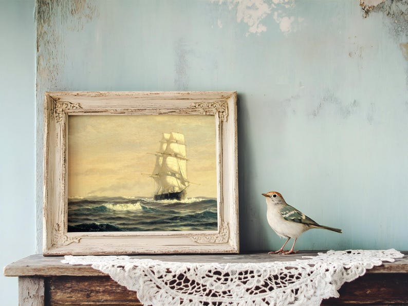 A framed painting of a tall sailing ship on the ocean, with billowing sails and waves, placed on a wooden surface. To the right of the painting, a small bird with white, gray, and black feathers and a hint of orange on its head is perched on a white lace doily, against a distressed, pale blue wall background.