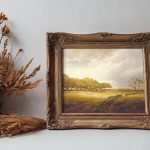 A rustic painting in an ornate frame shows a serene, sunlit landscape with trees and a path. Dried flowers in a basket are nearby, adding warmth.