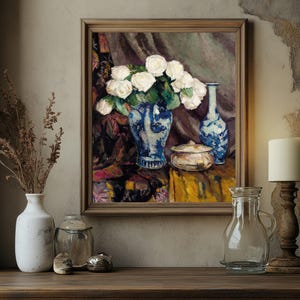 Framed painting of white roses in a blue vase on a textured wall. Surrounding objects include a lit candle, a clear glass bottle, and ceramic pots. Warm, cozy ambiance.
