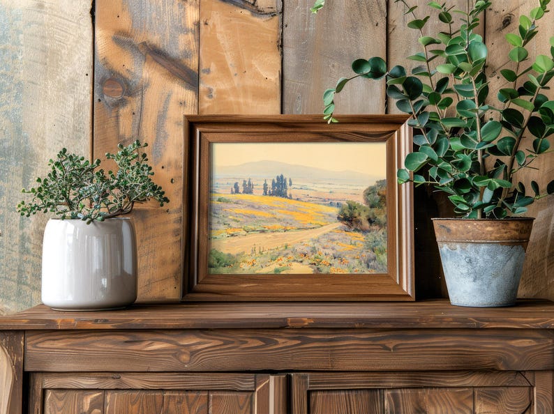 Framed landscape painting on wooden shelf, flanked by two potted plants. Rustic, natural vibe with a serene, artistic atmosphere.