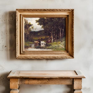 A rustic wooden table stands against a textured, aged wall with a framed pastoral painting above, depicting cattle by a stream under lush trees.