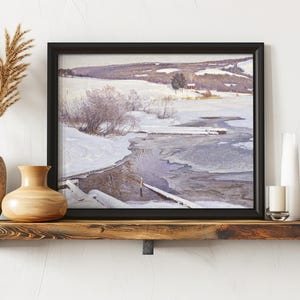 A framed winter landscape painting of a snowy countryside with a river sits on a wooden shelf. Nearby are decorative vases and candles, creating a cozy, serene atmosphere.