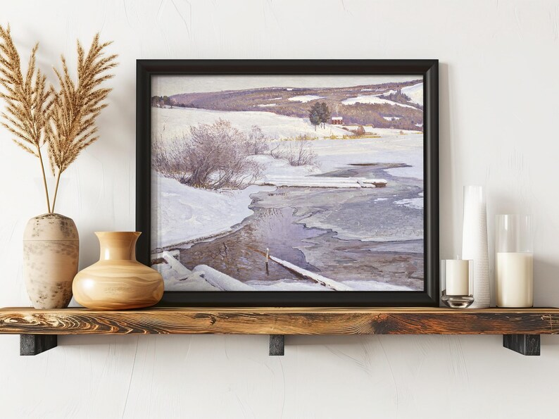 A framed winter landscape painting of a snowy countryside with a river sits on a wooden shelf. Nearby are decorative vases and candles, creating a cozy, serene atmosphere.