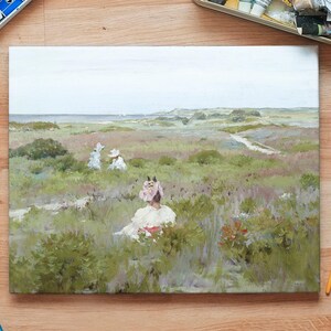 A landscape painting on a wooden table shows women in vintage dresses sitting in a lush, green field. Art supplies like paints and brushes surround the canvas.