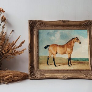 A vintage gold-framed painting of a horse on a sandy landscape is displayed on a shelf. Beside it, a rustic basket holds dried wildflowers.