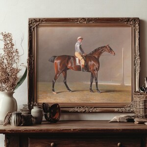 A framed painting of a jockey on a brown horse is displayed on a wooden table. The table also holds art supplies, a vase with dried flowers, and brushes.