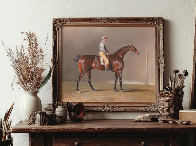 A framed painting of a jockey on a brown horse is displayed on a wooden table. The table also holds art supplies, a vase with dried flowers, and brushes.