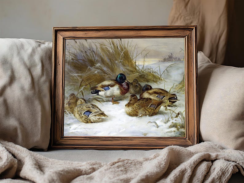 A framed painting of ducks resting on snow in a winter landscape with grass. The wooden frame sits on a cozy beige couch, evoking warmth.