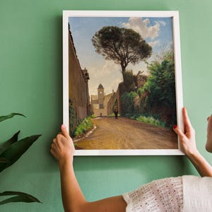 A framed painting depicting a street scene in a European village. The painting features a narrow street lined with buildings, a tall tree in the foreground, and a person walking in the distance.