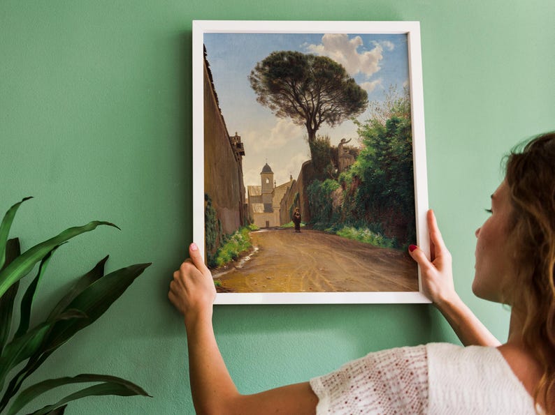 A framed painting depicting a street scene in a European village. The painting features a narrow street lined with buildings, a tall tree in the foreground, and a person walking in the distance.