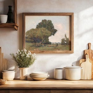 A cozy kitchen or dining area with a framed landscape painting of a large tree and open field hanging on a white textured wall. Below the painting, there is a wooden countertop with a potted plant with white flowers, a stack of white plates, and various white ceramic containers, including a lidded jar and a casserole dish. To the right, there are two wooden cutting boards, one leaning against the wall and the other standing upright.