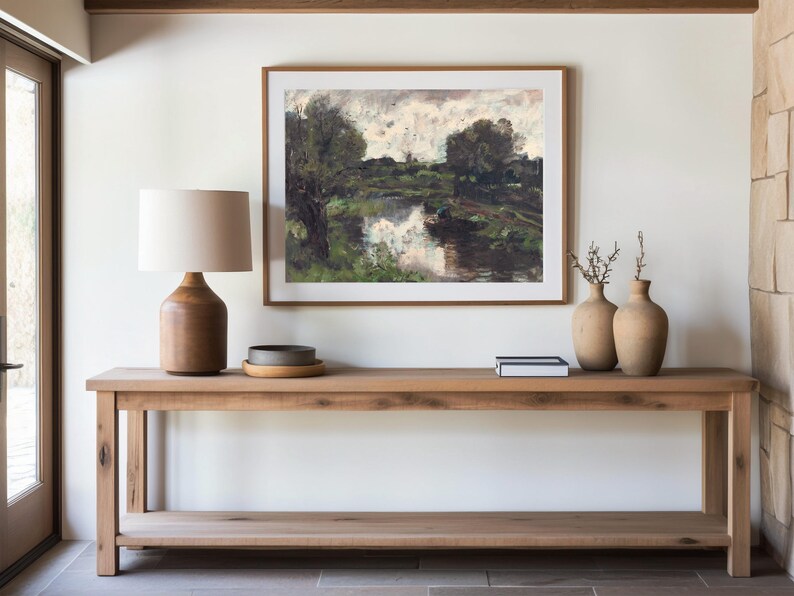 A minimalist room with a wooden console table. It holds a ceramic lamp and vases. Above, a framed landscape painting adds a serene touch.