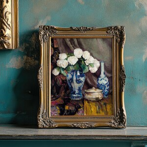 Framed painting of white roses in a blue vase on a textured teal wall. The ornate, gold frame complements the rustic and vintage ambiance.