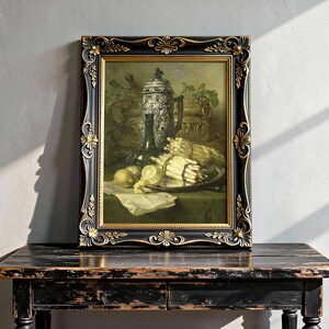A framed still life painting on a rustic wooden table shows a decorative jug, a wine bottle, and a platter with asparagus and lemons. Soft sunlight casts shadows on a textured wall.