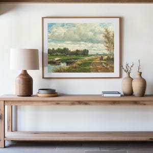 A serene interior features a wooden console table topped with a lamp and vases. Above, a landscape painting depicts a river and path under a cloudy sky.
