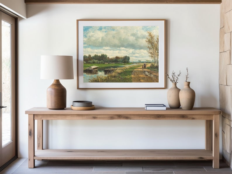 A serene interior features a wooden console table topped with a lamp and vases. Above, a landscape painting depicts a river and path under a cloudy sky.