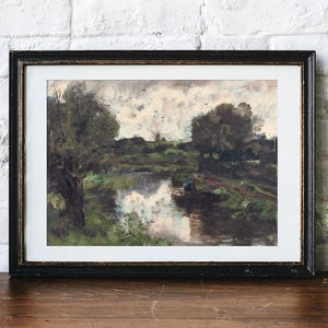 Framed landscape painting on a wooden table, depicting a serene river with lush greenery and trees. Cactus in a pot on the right adds a cozy feel.