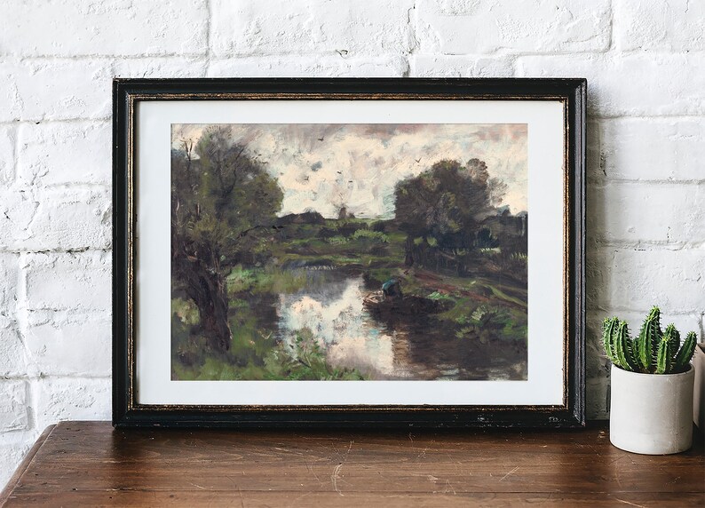 Framed landscape painting on a wooden table, depicting a serene river with lush greenery and trees. Cactus in a pot on the right adds a cozy feel.