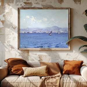 Cozy living room with a beige sofa, rust-colored cushions, and a blanket. A sea landscape painting hangs on a textured wall, evoking tranquility.