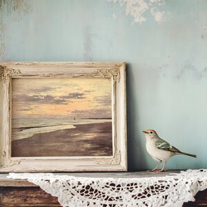 A small bird perches on an old wooden table draped with lace. Beside it, a rustic frame holds a painting of an ocean sunset, set against a pale blue wall.