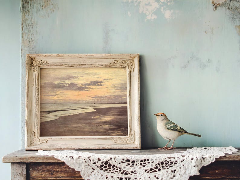 A small bird perches on an old wooden table draped with lace. Beside it, a rustic frame holds a painting of an ocean sunset, set against a pale blue wall.