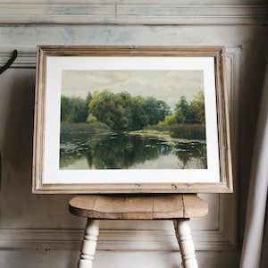 A framed landscape painting depicting a serene lake scene with lush green trees and reflections in the water. The artwork is encased in a light brown wooden frame and rests on a small, rustic wooden stool.