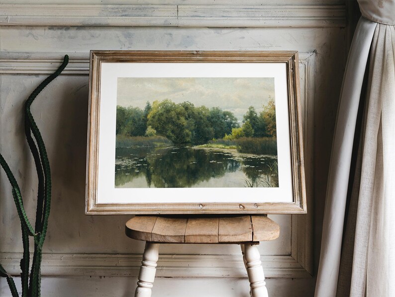A framed landscape painting depicting a serene lake scene with lush green trees and reflections in the water. The artwork is encased in a light brown wooden frame and rests on a small, rustic wooden stool.