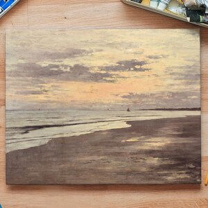 Painting of a serene sunset over the ocean, with a sandy beach in the foreground. Art supplies, including brushes and paints, surround the artwork.