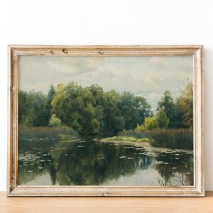 A framed landscape painting depicts a tranquil waterscape. Lush green trees and foliage line the water's edge, reflecting in the dark water. The painting has a vintage aesthetic with a light-colored, distressed wooden frame.