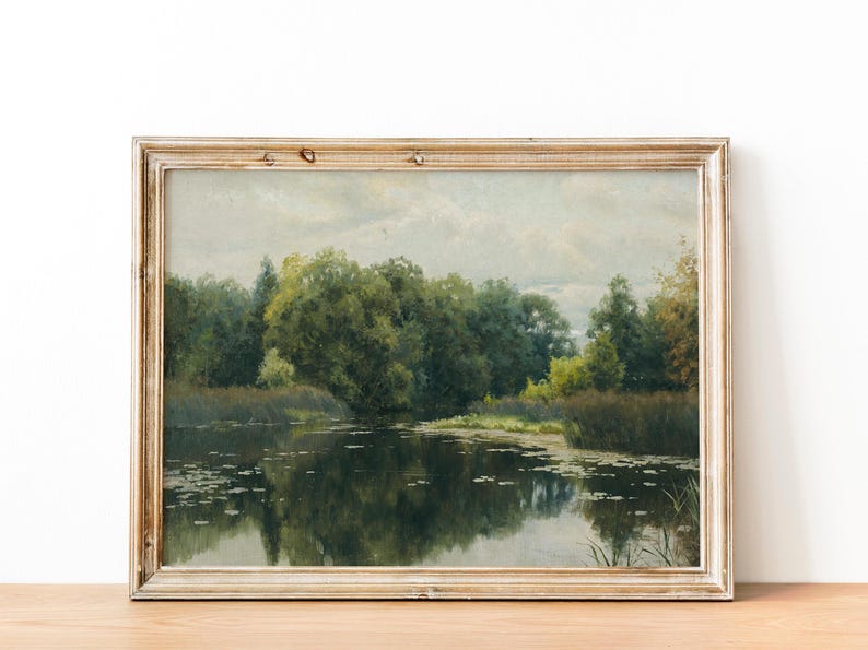 A framed landscape painting depicts a tranquil waterscape. Lush green trees and foliage line the water's edge, reflecting in the dark water. The painting has a vintage aesthetic with a light-colored, distressed wooden frame.