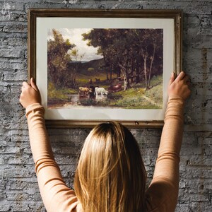 A woman hangs a framed landscape painting on a brick wall. The painting depicts a rural scene with trees, a stream, and cows.