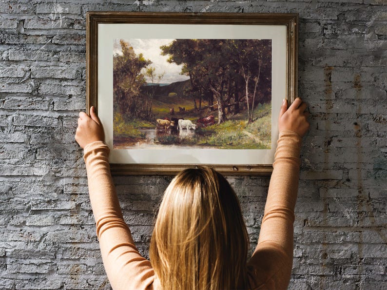 A woman hangs a framed landscape painting on a brick wall. The painting depicts a rural scene with trees, a stream, and cows.