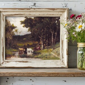 A rustic, framed painting of cows by a stream is on a wooden shelf. Beside it, a Mason jar holds wildflowers, adding a touch of nature.