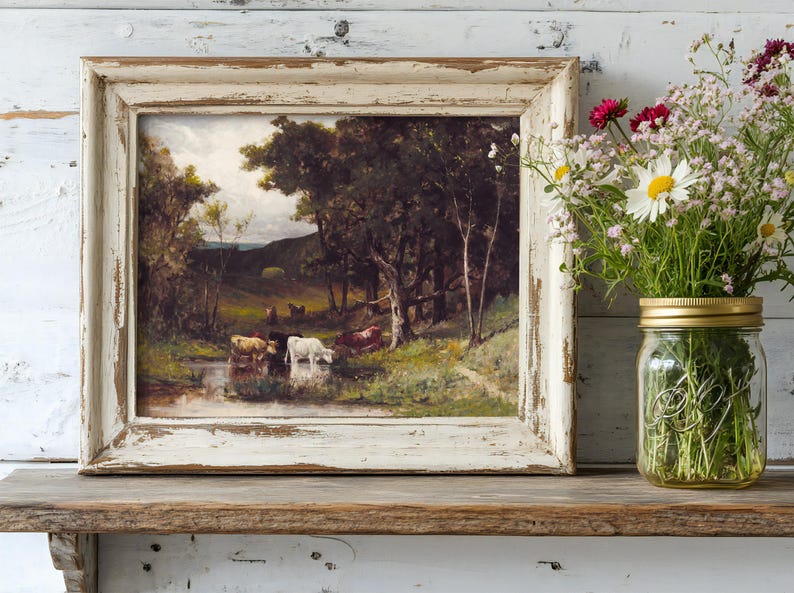 A rustic, framed painting of cows by a stream is on a wooden shelf. Beside it, a Mason jar holds wildflowers, adding a touch of nature.
