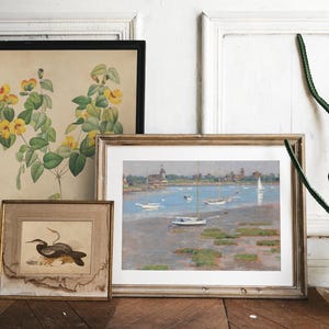 Three framed pictures hanging on a white wall. The picture on the left is a botanical illustration of yellow flowers with green leaves. The middle picture is a painting of a seascape with sailboats and a lighthouse. The picture on the right is a black and white illustration of a bird.