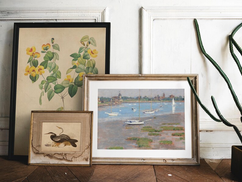 Three framed pictures hanging on a white wall. The picture on the left is a botanical illustration of yellow flowers with green leaves. The middle picture is a painting of a seascape with sailboats and a lighthouse. The picture on the right is a black and white illustration of a bird.
