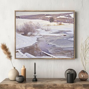 A winter landscape painting hangs on a textured wall above a wooden table. The scene shows a snowy field with a partially frozen stream. The table displays neutral-toned vases and a candle, providing a calm, rustic feel.