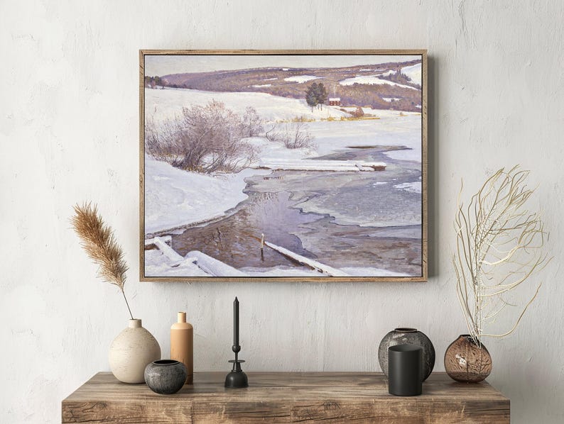 A winter landscape painting hangs on a textured wall above a wooden table. The scene shows a snowy field with a partially frozen stream. The table displays neutral-toned vases and a candle, providing a calm, rustic feel.