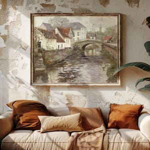 Cozy living room with a white sofa adorned with brown and beige cushions. A large painting of a scenic village and bridge above, flanked by lush plants.
